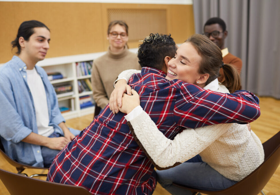 Portrait of two young women hugging in support group meeting, both smiling happily, copy space